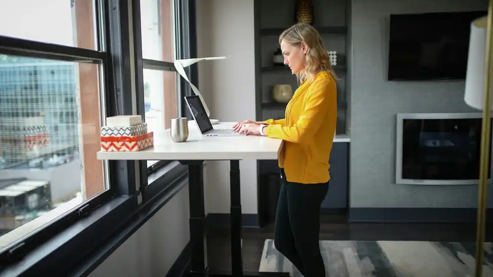 Mujer trabajando de pie con portátil sobre un escritorio elevable eléctrico en un entorno luminoso y moderno con vistas exteriores.