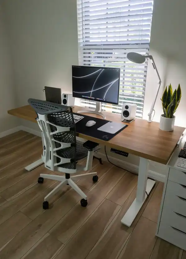 Setup minimalista con marco de escritorio elevable blanco y tablero de madera, ideal para home office con monitor, altavoces y silla ergonómica.