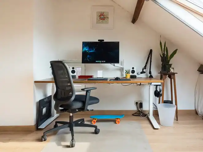 Habitación abuhardillada con estructura de escritorio elevable blanca y setup completo con monitor, altavoces y accesorios, sobre un marco ergonómico ajustable.