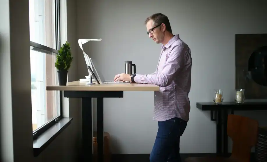 Hombre trabajando de pie con portátil sobre un escritorio elevable manual de madera y estructura negra, junto a una ventana en un entorno moderno de teletrabajo.
